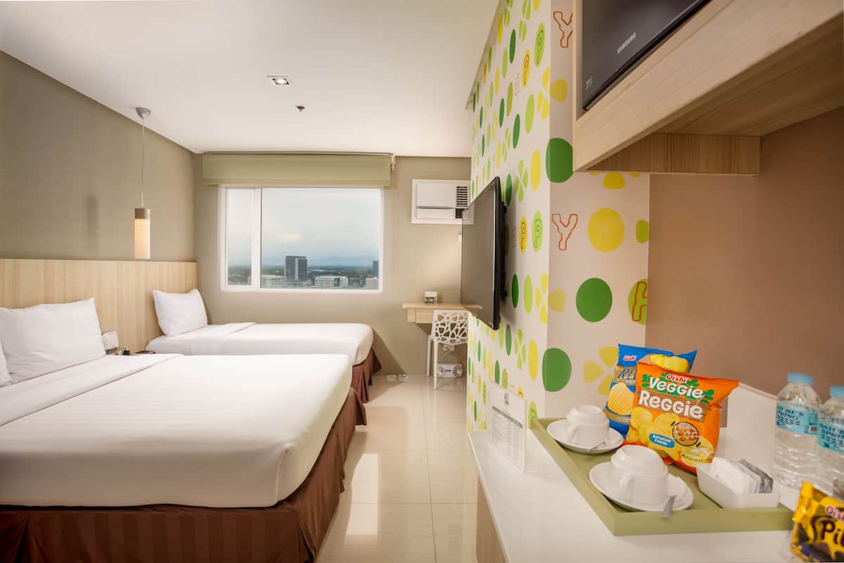 Ein Hotelzimmer mit zwei Betten, einem Fernseher, einem Fenster mit Blick auf die Stadt, einem Schreibtisch mit Stuhl und einer Snackbar mit Veggie Reggie Chips.