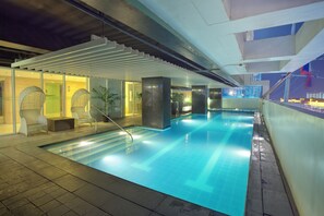 A modern indoor swimming pool with clear blue water, surrounded by lounge chairs and potted plants.