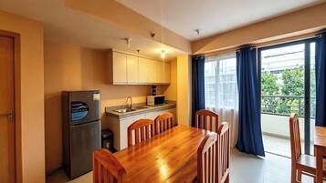 A compact kitchen with a refrigerator, microwave, and sink, a wooden dining table with chairs, and a large window with curtains.