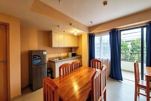 A compact kitchen with a refrigerator, microwave, and sink, a wooden dining table with chairs, and a large window with curtains.