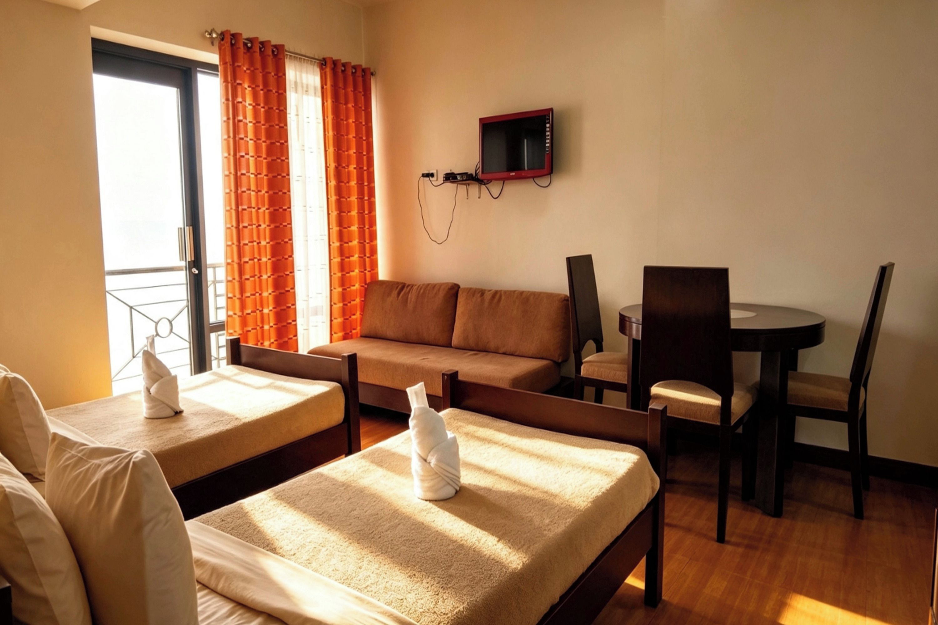 A hotel room with a bed, a sofa, a dining table with chairs, a TV mounted on the wall, and a window with curtains.