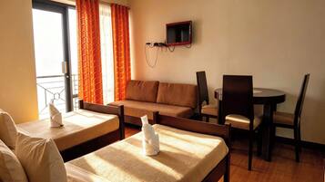 A hotel room with a bed, a sofa, a dining table with chairs, a TV mounted on the wall, and a window with curtains.
