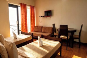 Una habitación de hotel con cama, sofá, mesa de comedor con sillas, televisor montado en la pared y ventana con cortinas.
