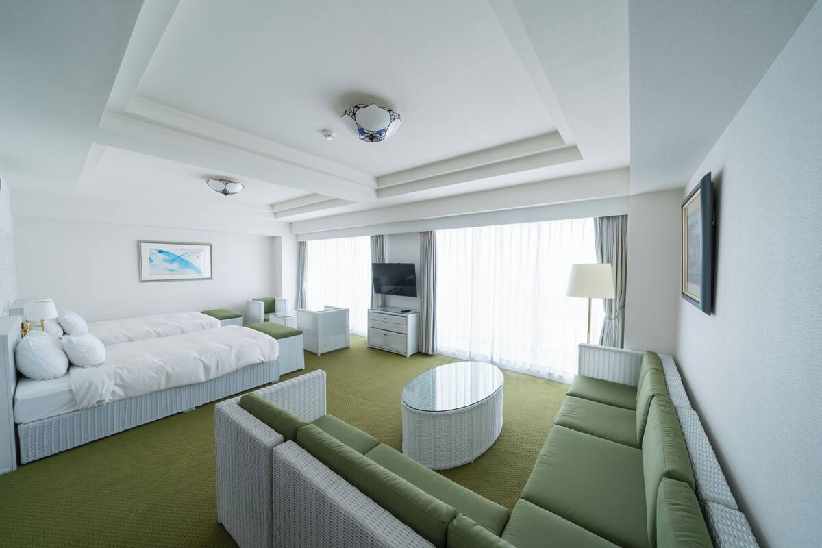 Une chambre d’hôtel avec un lit, un canapé vert, une table basse ronde, une télévision et un tableau au mur.