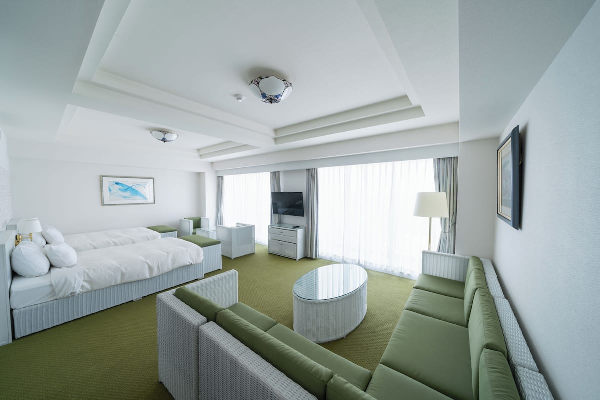 Hotellihuone, jossa on sänky, vihreä sohva, pyöreä sohvapöytä, televisio ja seinällä taulu.