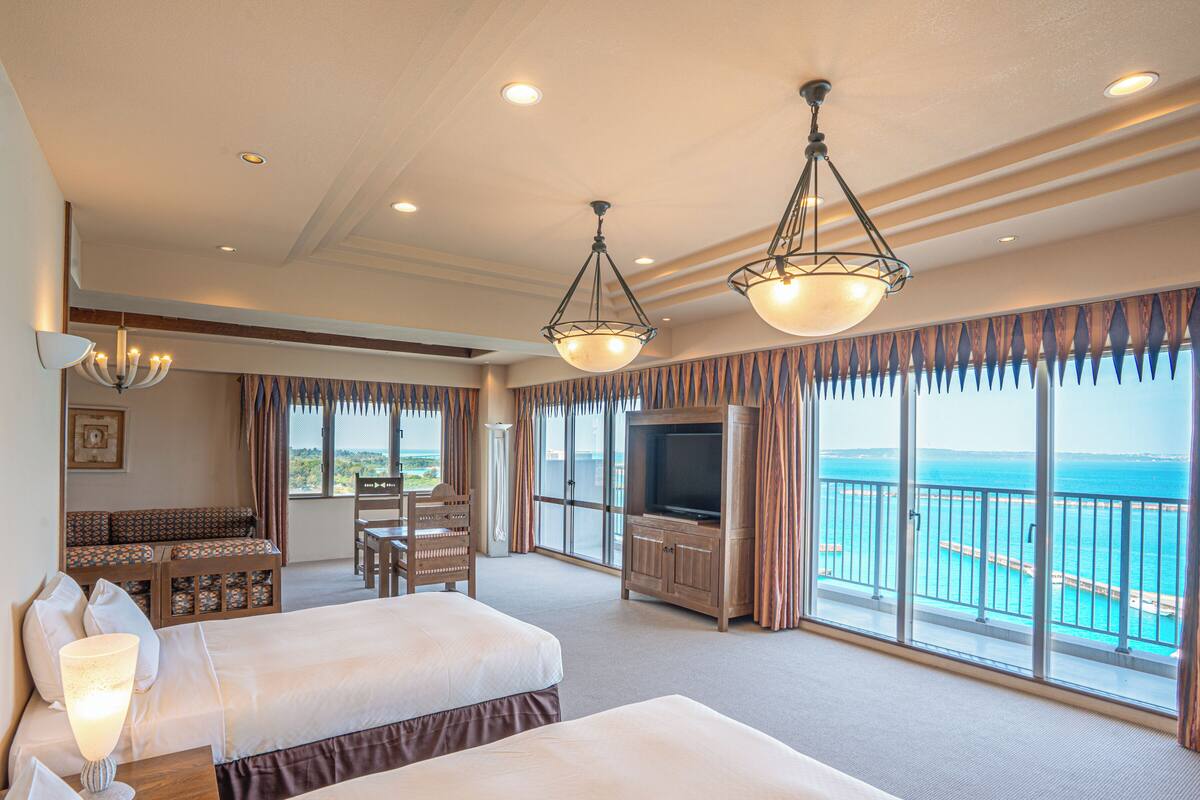 Une chambre d’hôtel avec deux lits, une télévision, un balcon avec vue sur la mer et un coin repas avec une table et des chaises.