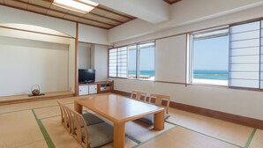 Ein traditioneller japanischer Raum mit Tatami-Boden, einem Holztisch mit Stühlen, einem Fernseher und großen Fenstern mit Meerblick.