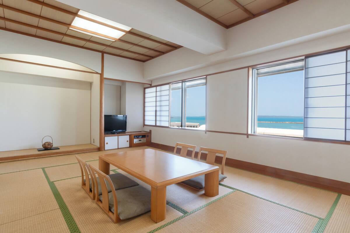 Une pièce de style japonais traditionnel, avec un sol recouvert de tatamis, une table en bois entourée de chaises, un téléviseur et de grandes fenêtres offrant une vue sur la mer.