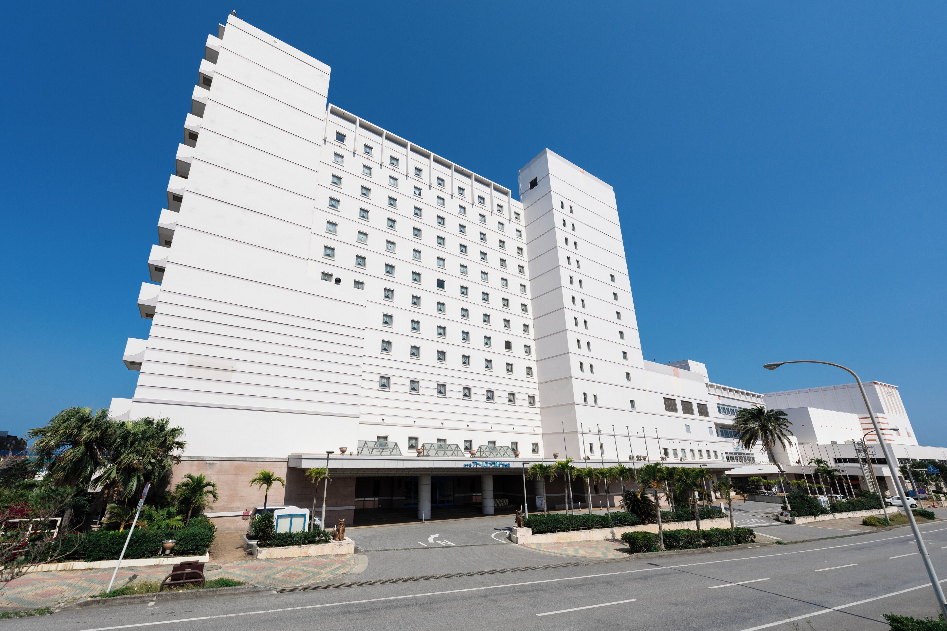 Un hotel grande de varios pisos con diseño moderno, que presenta una entrada destacada con zona cubierta y palmeras.