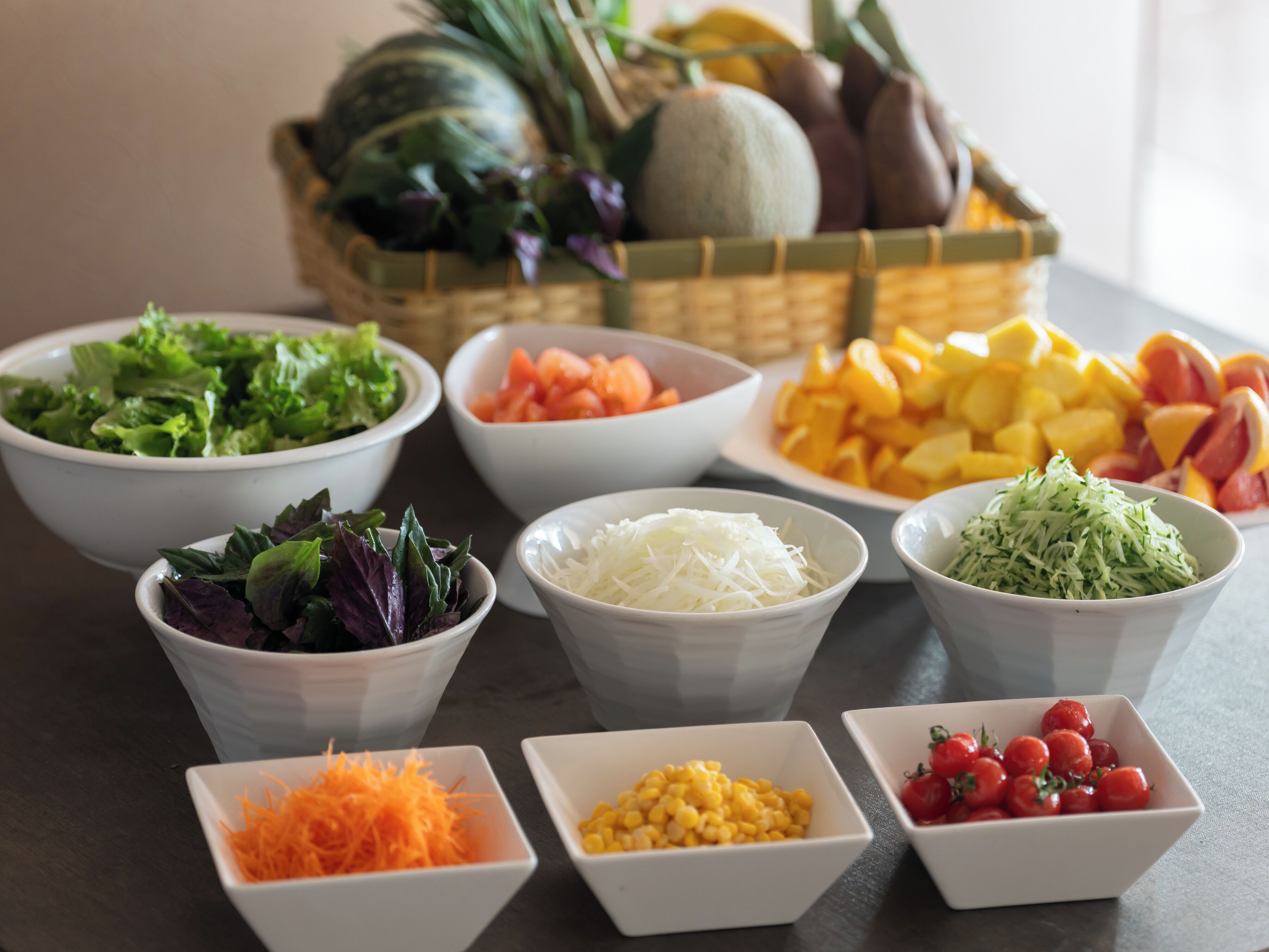 A variety of fresh vegetables and fruits in bowls, including lettuce, carrots, corn, and tomatoes, arranged on a table.