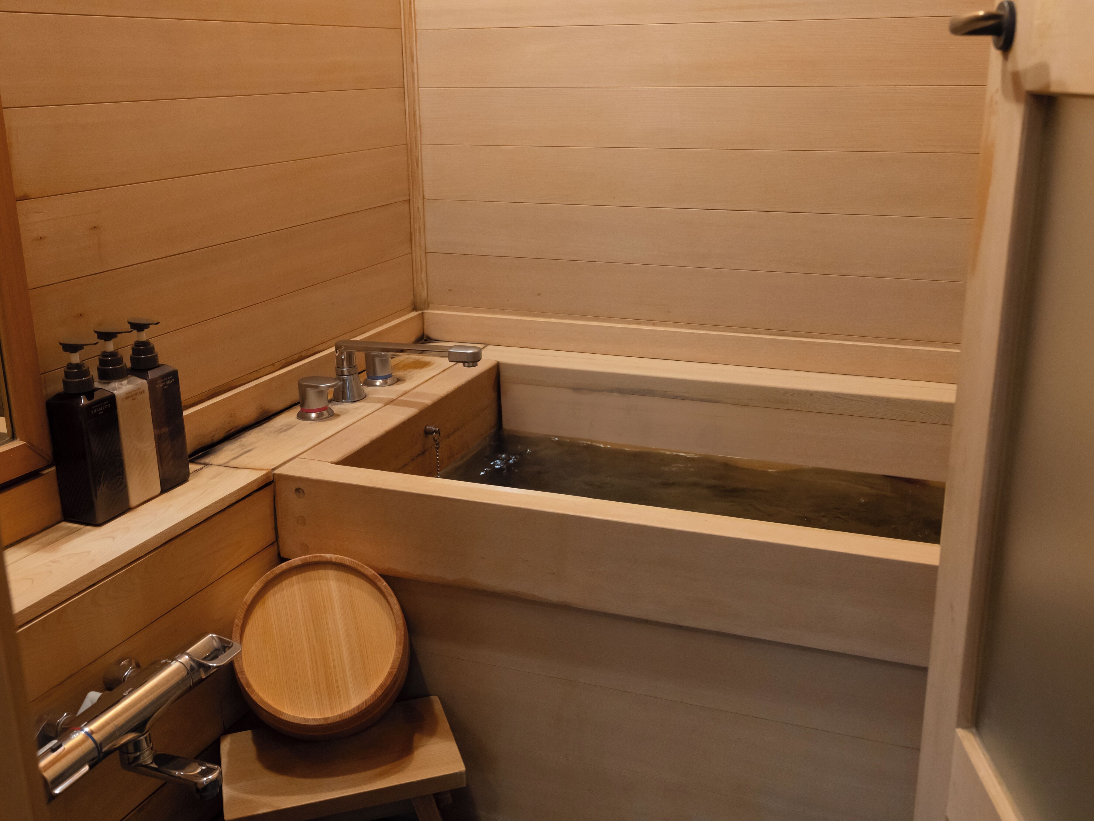 A wooden-paneled bathroom with a bathtub, a wooden basin, and three bottles of toiletries on a shelf.