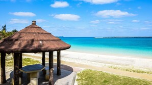 Ein Strandpavillon mit Reetdach und Tisch, mit Blick auf das klare, blaue Meer und den Sandstrand.