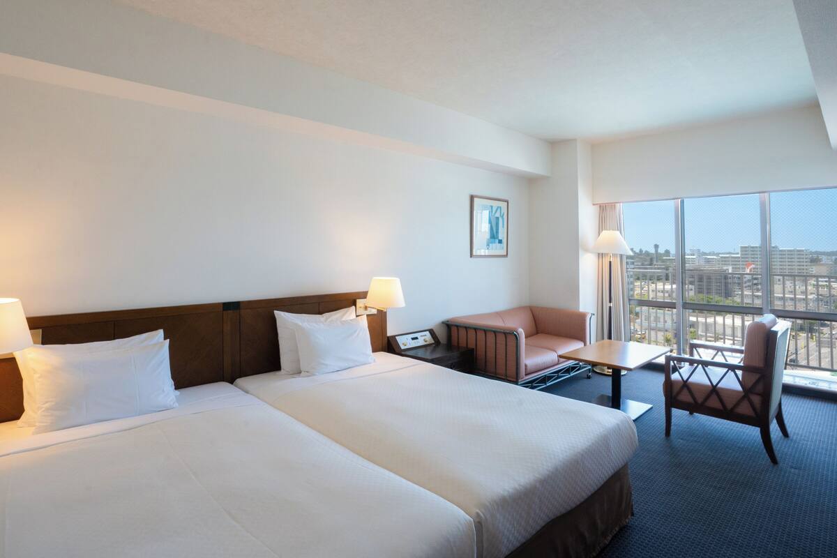 Une chambre d’hôtel avec deux lits, un bureau, une chaise et une grande fenêtre donnant sur la ville.