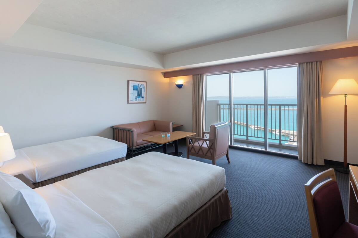 Une chambre d’hôtel avec deux lits, un bureau, une chaise, une vue sur la mer et un tableau au mur.
