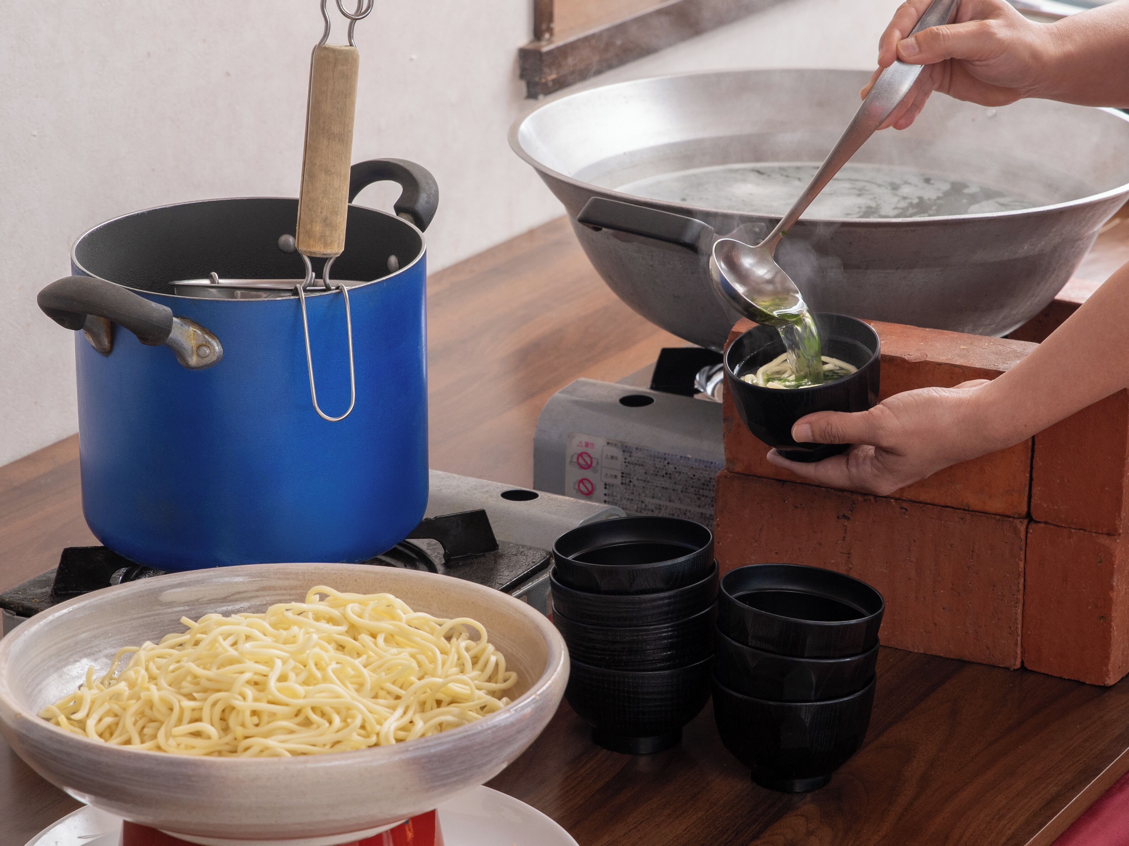 A kitchen with a blue pot on a stove, a large metal bowl, and a person cooking.