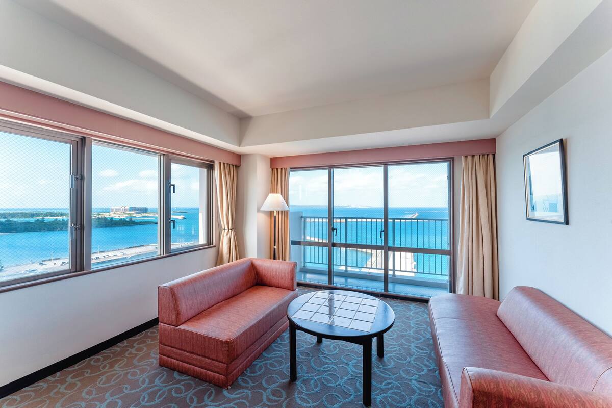 Une chambre d’hôtel avec un balcon, deux canapés, une petite table et une vue sur la mer.
