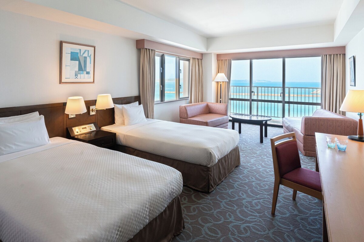 A hotel room with two beds, a desk, a chair, and a view of the sea.