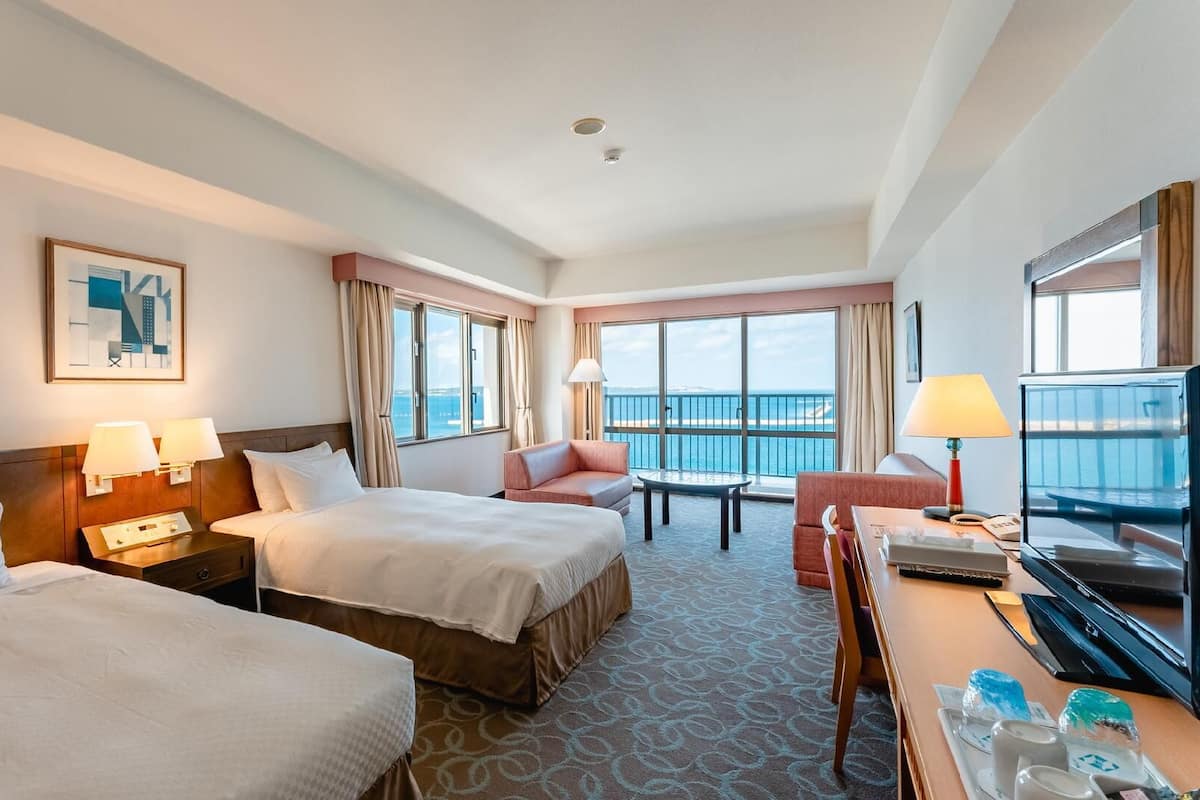 A hotel room with two beds, a desk with a computer, a chair, a small table, and a view of the ocean.