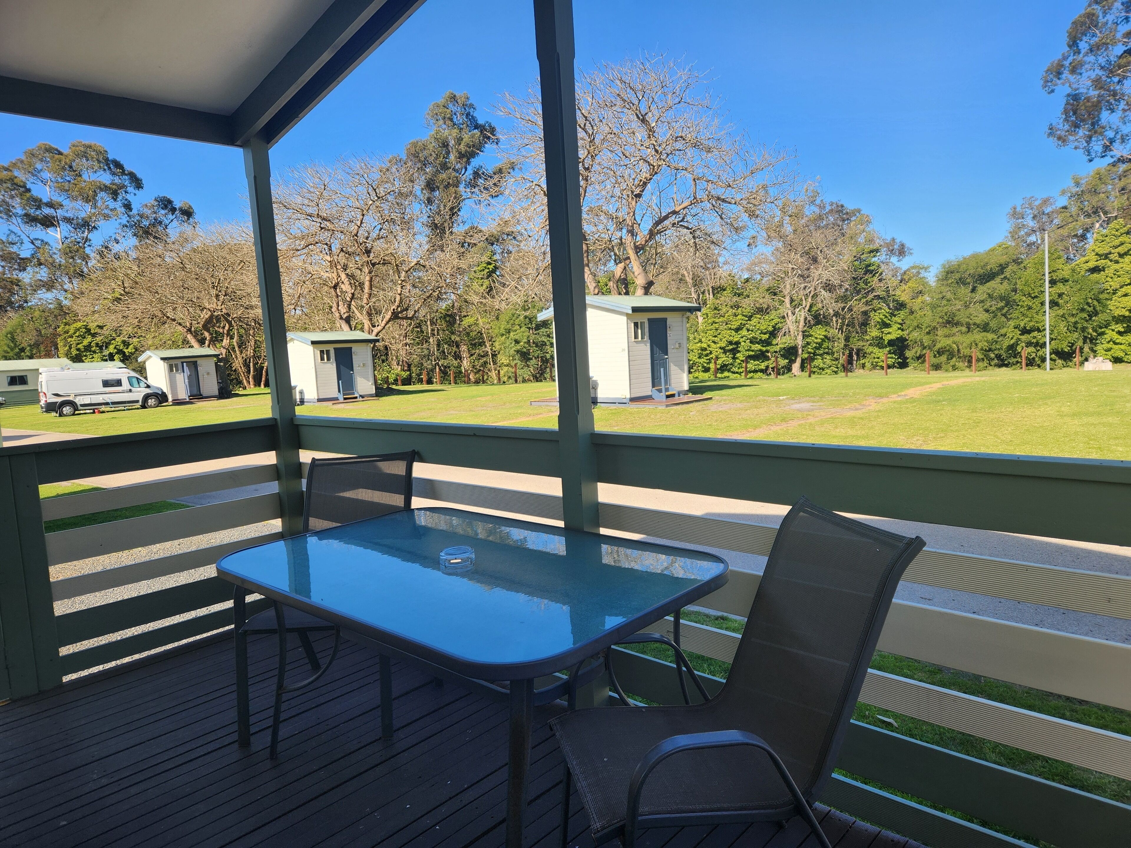A covered outdoor area with a table and chairs, a small building, and a grassy field with trees in the background.