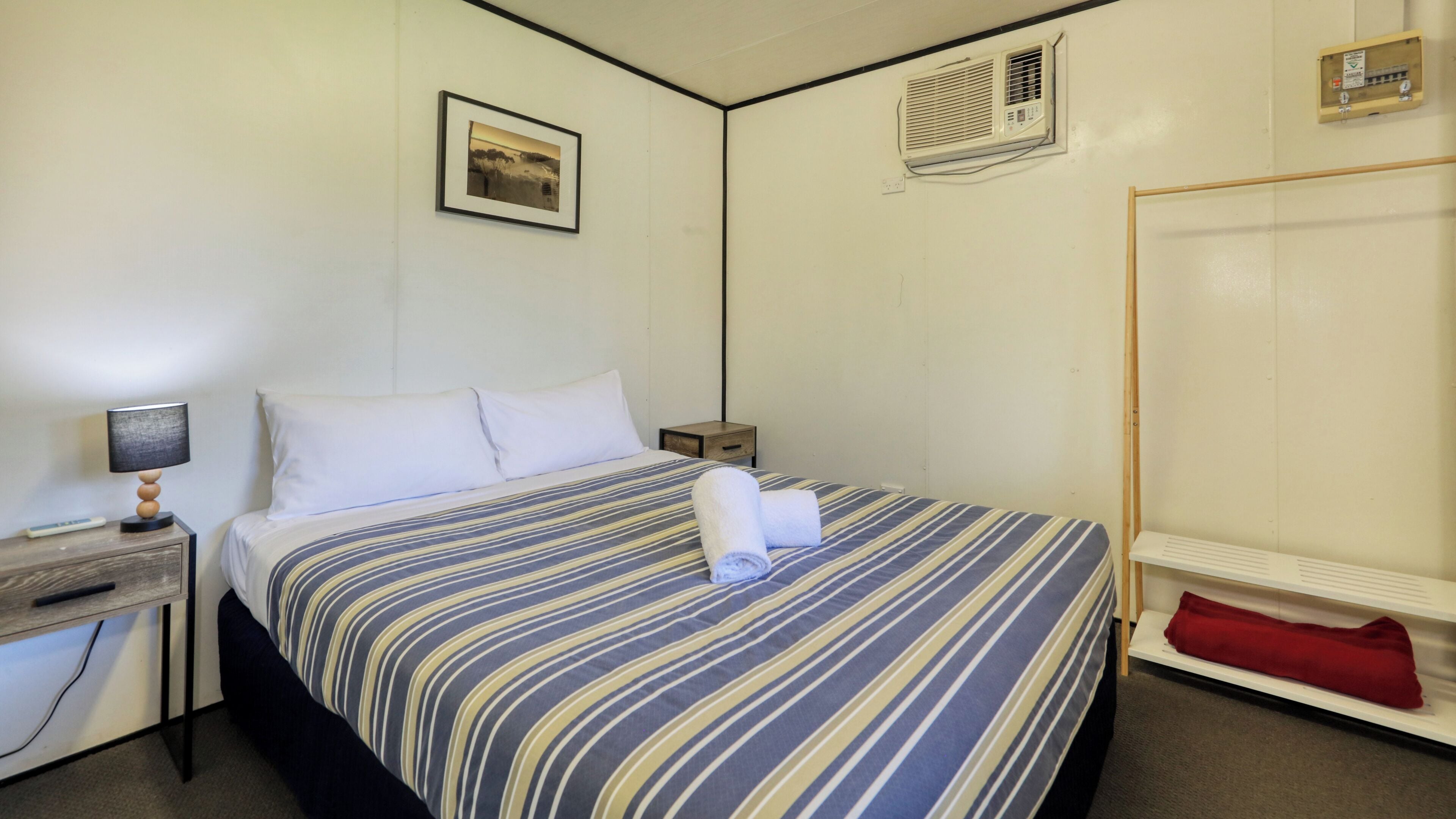 A single bed with a striped bedspread, a bedside table with a lamp, a small wooden nightstand, a framed picture on the wall, an air conditioning unit, and a wardrobe.