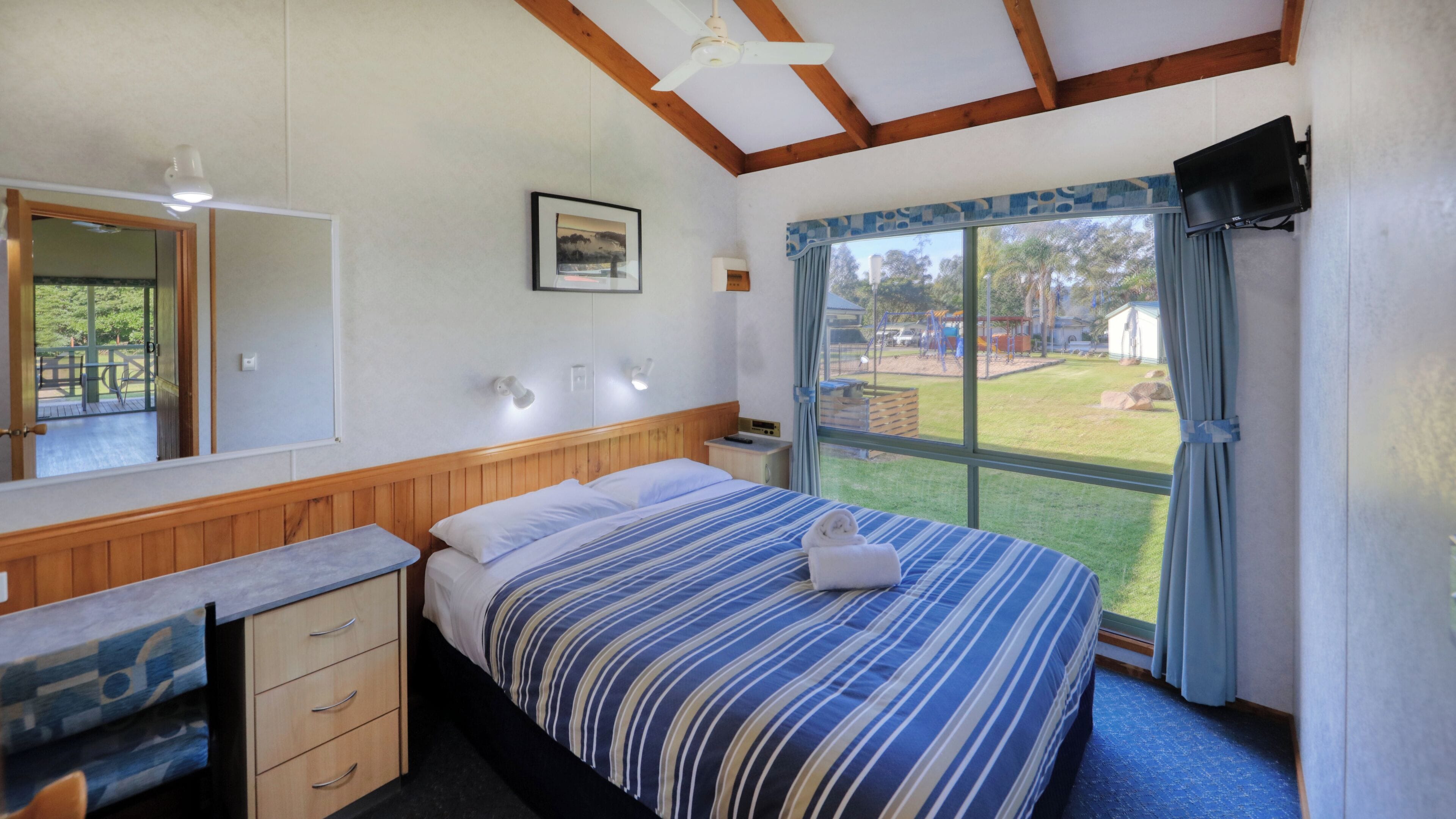 A bedroom with a bed, a dresser, a mirror, a window with curtains, and a television.