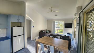 A compact kitchen with a refrigerator, a dining table with chairs, a ceiling fan, and a window with a view of greenery outside.