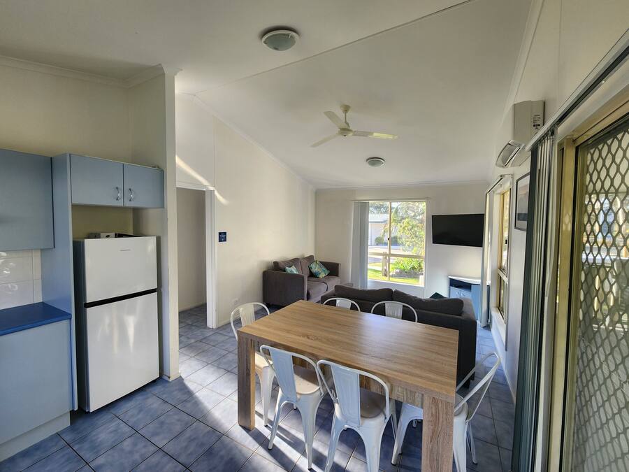 A compact kitchen with a refrigerator, a dining table with chairs, a ceiling fan, and a window with a view of greenery outside.