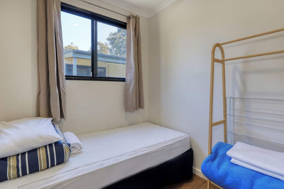 A small, single-bed room with a window, a wooden ladder, a blue towel, and a striped pillow.