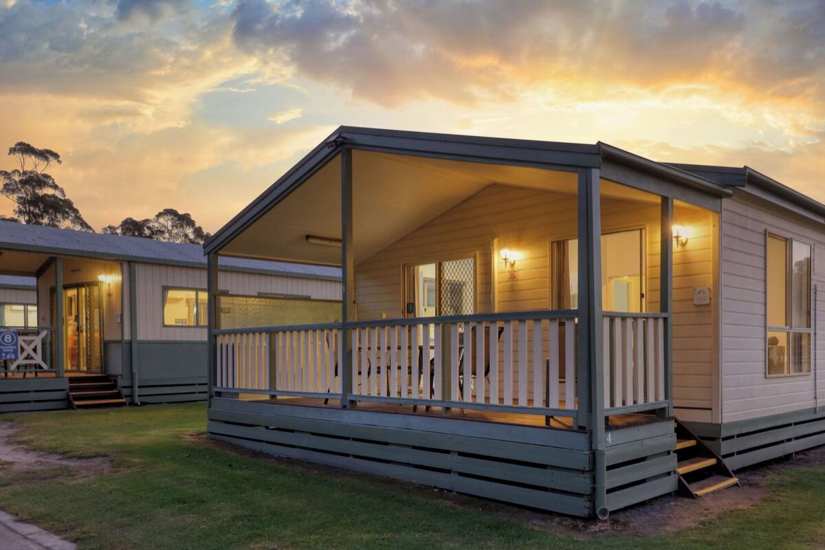 A small, single-story cabin with a porch, a fenced yard, and a sunset sky.