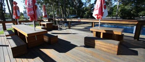 Una terraza de madera con mesas de picnic y sombrillas, con vista a un cuerpo de agua.