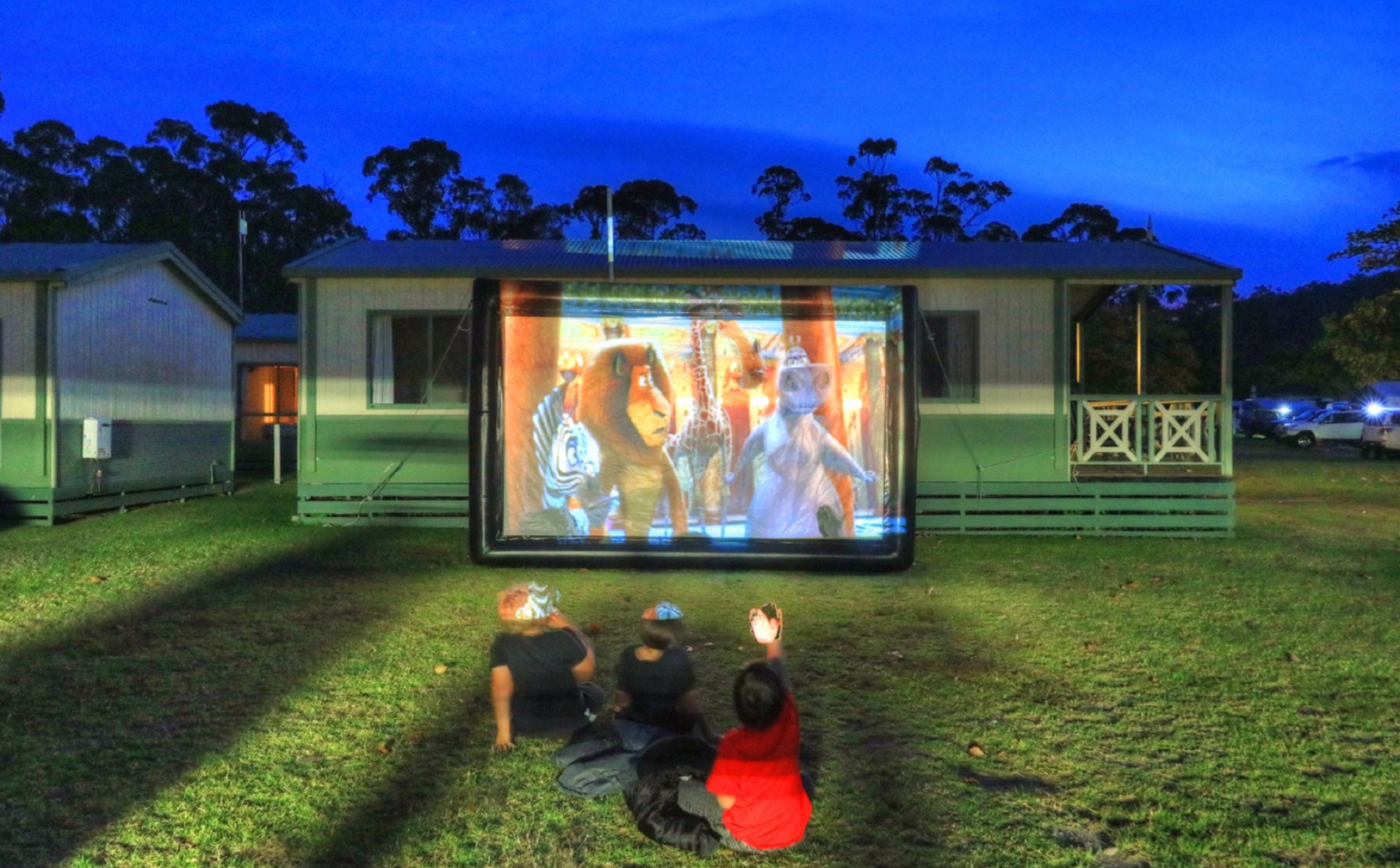 Una pantalla de cine portátil al aire libre mostrando personajes animados, montada en una zona de césped con un edificio al fondo.
