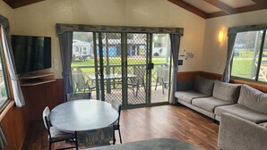Una habitación acogedora con un televisor grande, una mesa redonda, un sofá y un ventanal corredizo que da a un patio exterior con sillas y una parrilla.