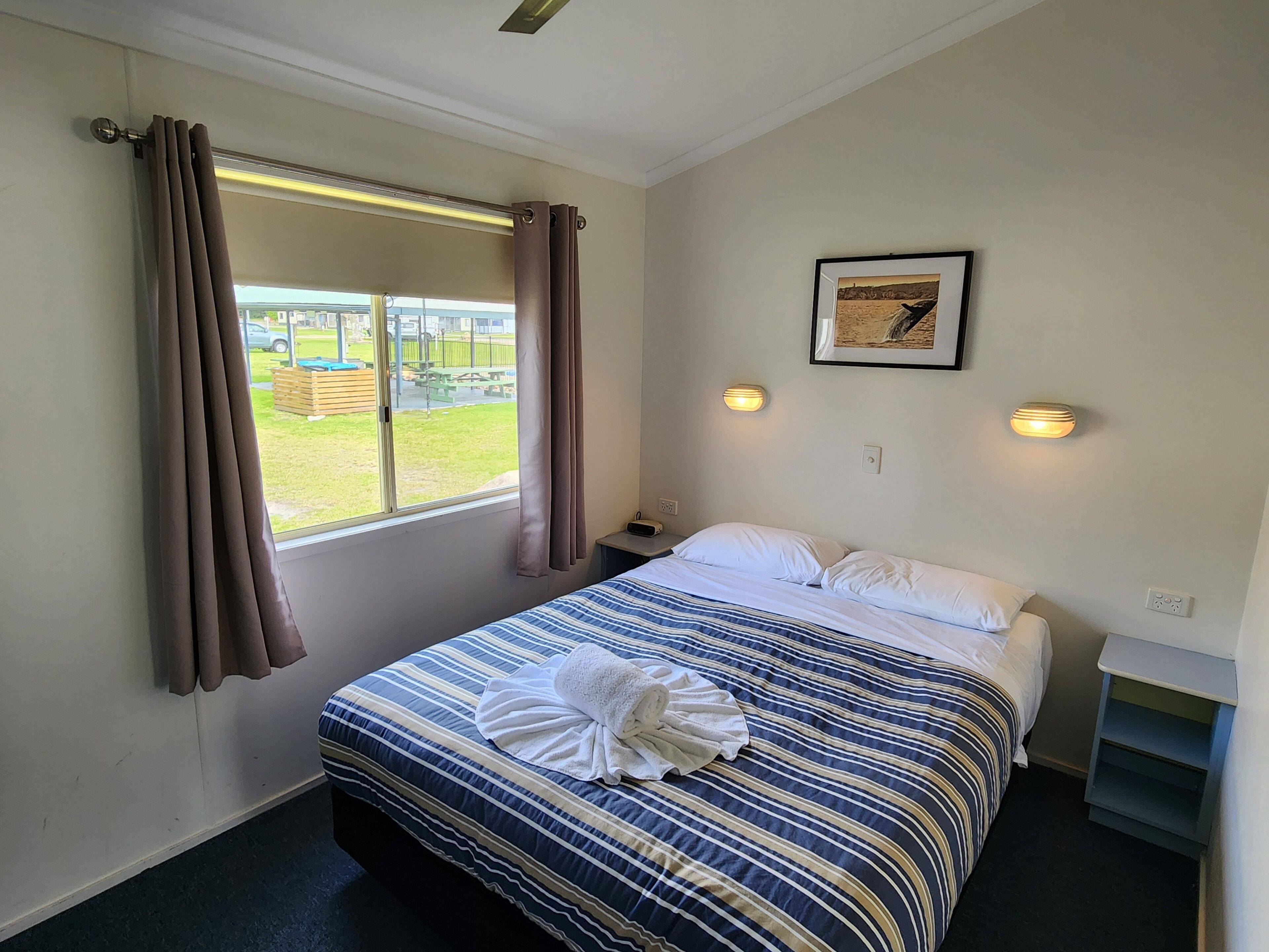 A hotel room with a bed, a window with curtains, a bedside table, a wall-mounted light, and a framed picture.