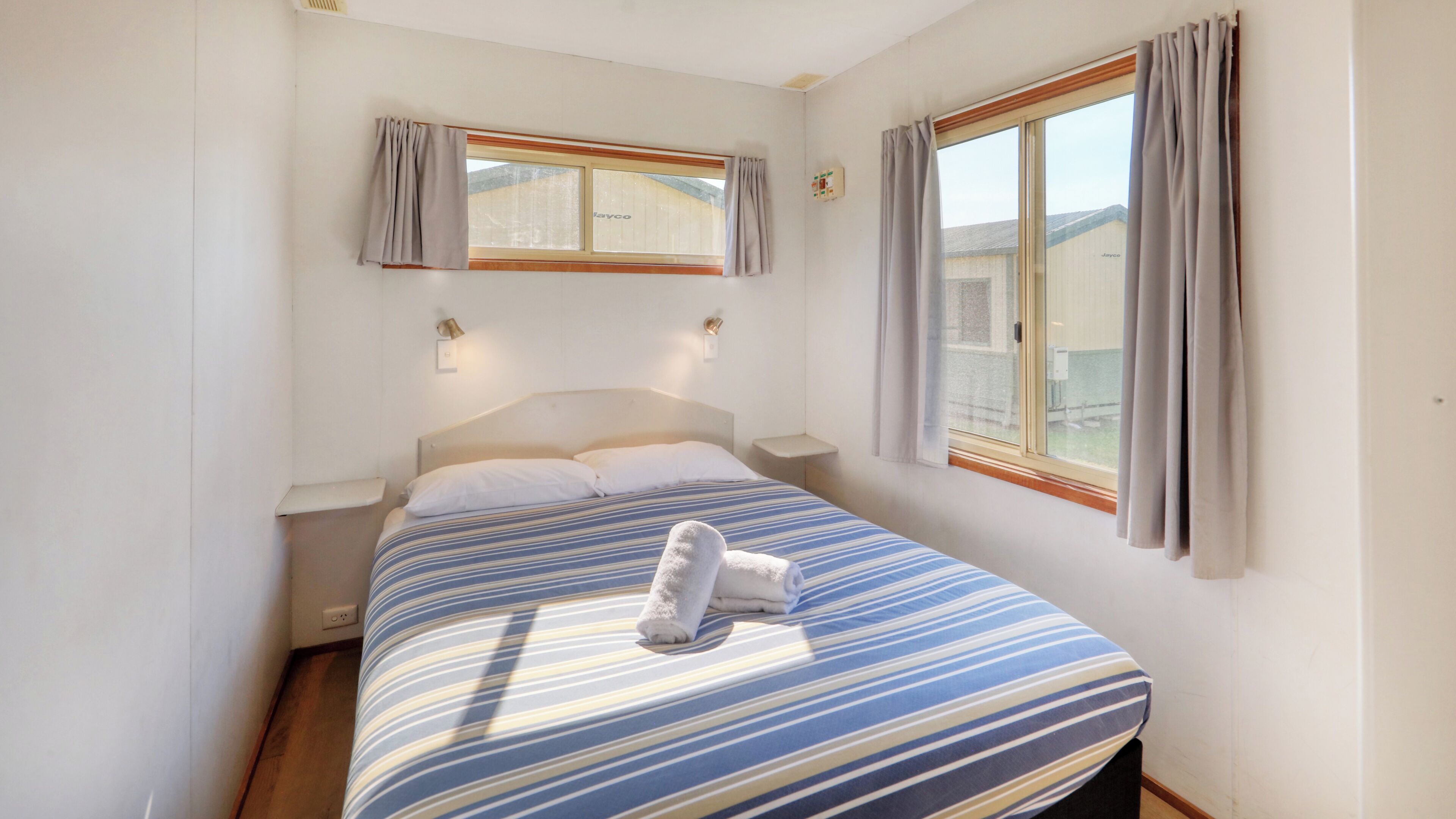 A single bed with a striped bedspread, two bedside tables, a nightstand, a window with curtains, and a wall-mounted light fixture.