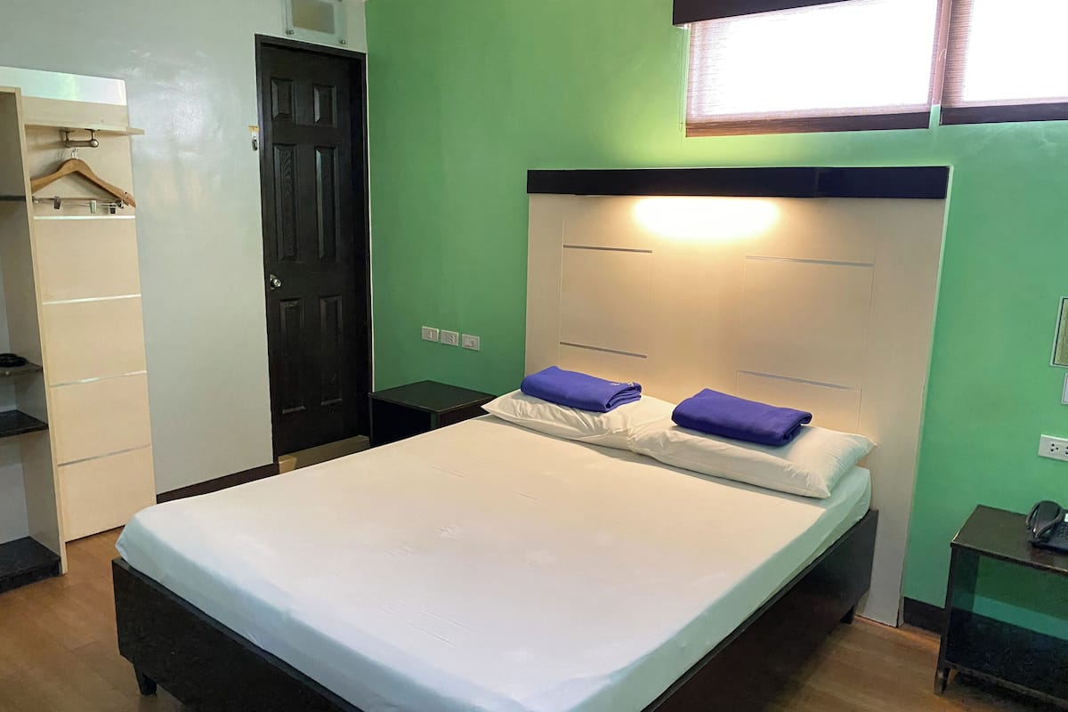 A single-bed room with a wooden headboard, a bedside table with a lamp, and a wardrobe with a hanging rack.