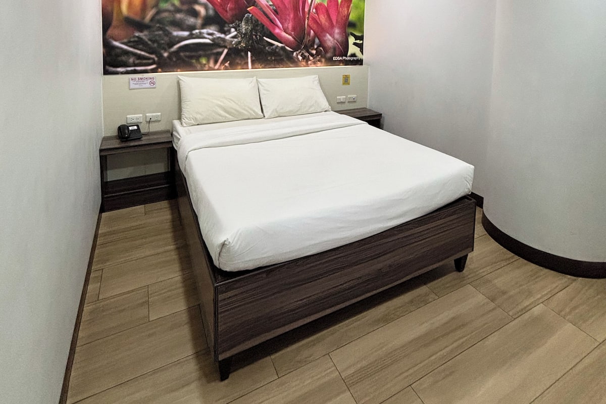 A neatly made bed in a hotel room with a wooden floor, a nightstand with a phone, and a 'No Smoking' sign.