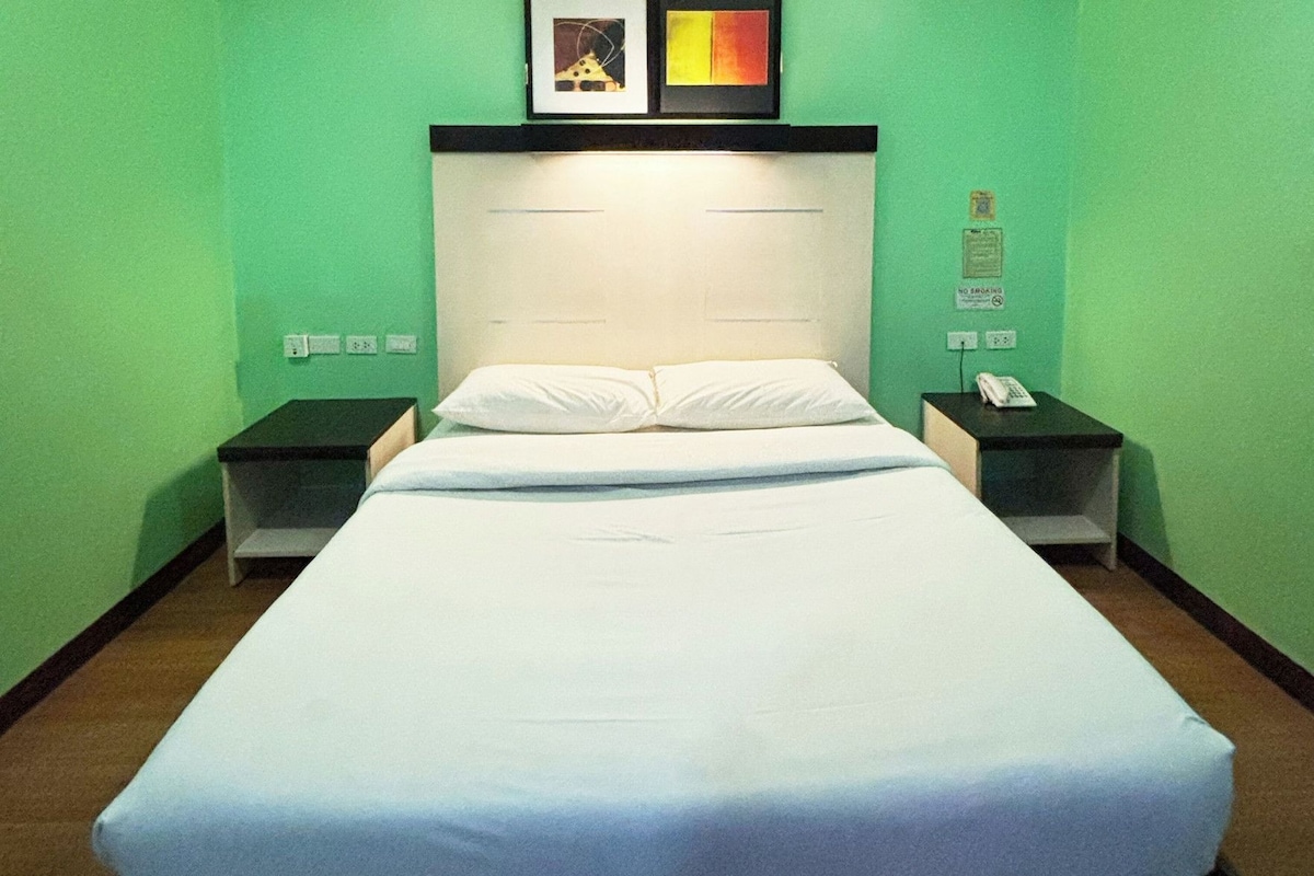 A hotel room with a single bed, two bedside tables, a nightstand with a phone, and a wall-mounted artwork above the bed.