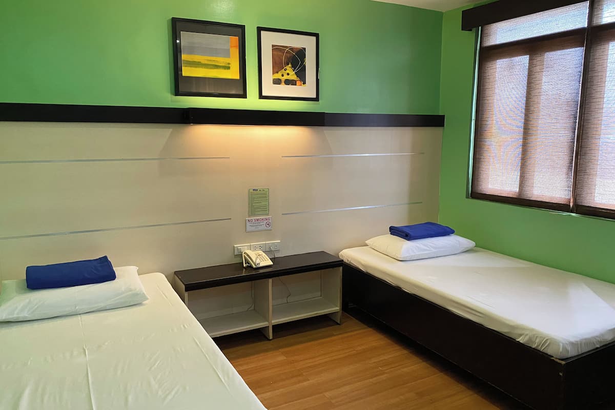 A room with two single beds, a small desk with a telephone, and a window with blinds.