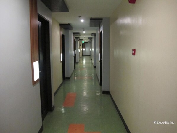 Un pasillo largo y estrecho con puertas a ambos lados, un suelo verde con marcas anaranjadas y una señal de salida de emergencia roja en la pared derecha.