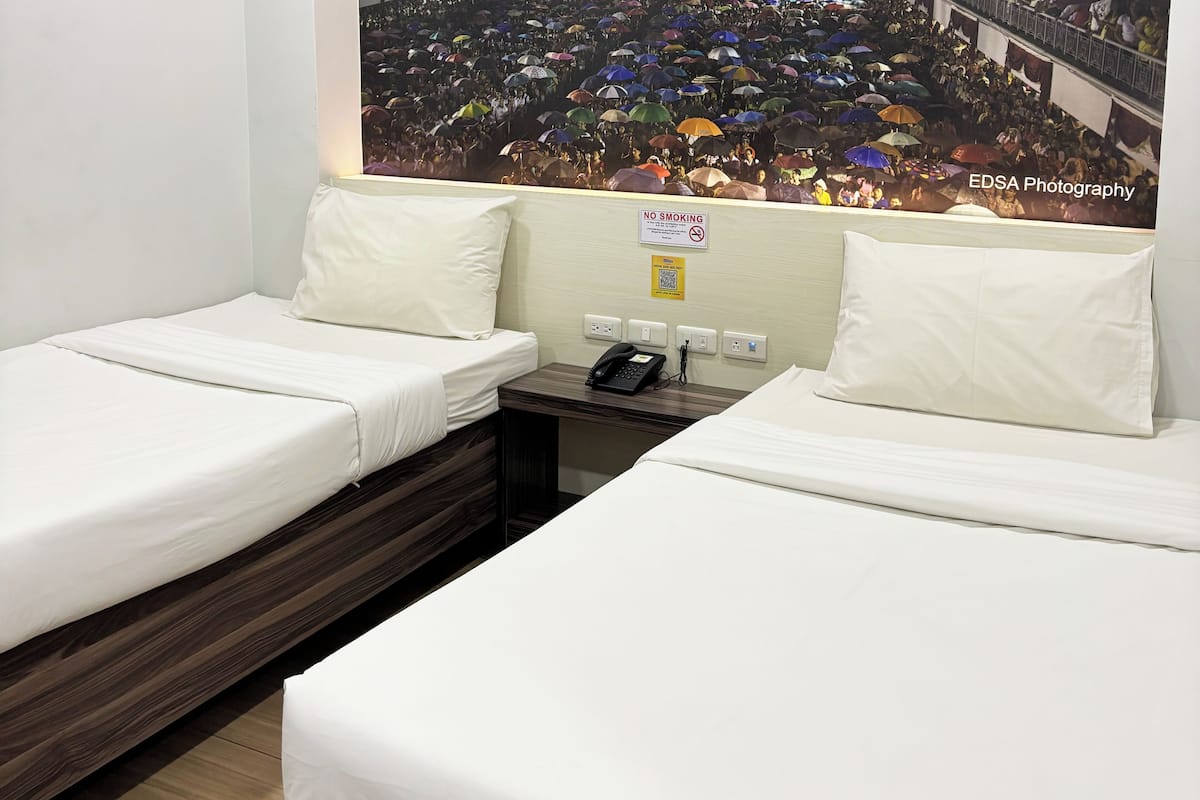 A hotel room with two single beds, a bedside table with a phone, and a wall-mounted sign indicating no smoking.