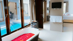 A modern hotel room with a large bathtub filled with rose petals, a bed with white linens, a television mounted on the wall, and a view of a swimming pool through the window. - Luxotic Private Villa and Resort (Nusa Dua)