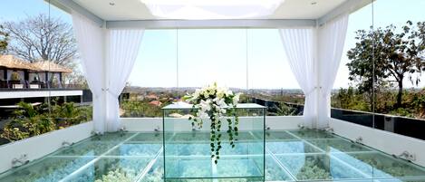 A glass-enclosed balcony with a clear view of the surrounding landscape, a glass table with a floral arrangement, and white curtains framing the windows.