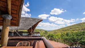 A wooden deck with a view of a lush green valley, a thatched roof structure, and a seating area with cushions.