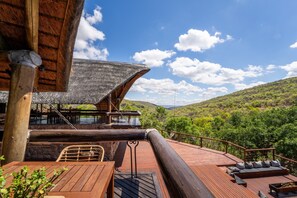 Une terrasse en bois offrant une vue sur une vallée verdoyante, une structure dotée d’un toit de chaume et un espace détente avec des coussins.