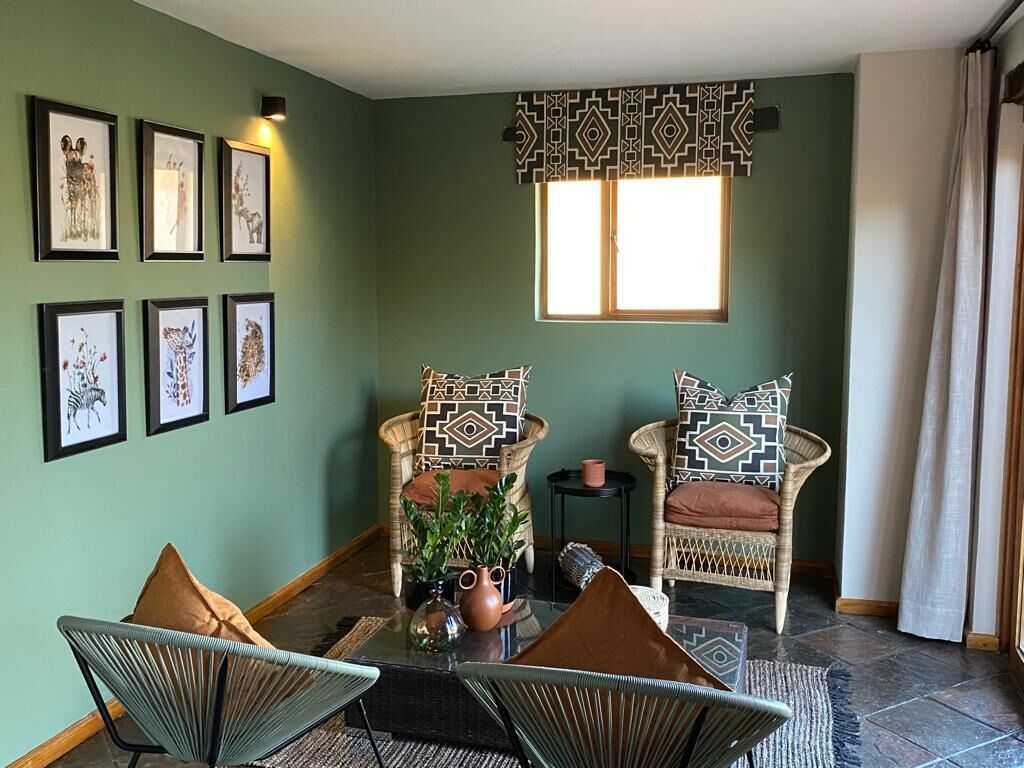 A room with a green wall, framed animal prints, wicker chairs with patterned cushions, a small table with a plant, and a window with a patterned valance.