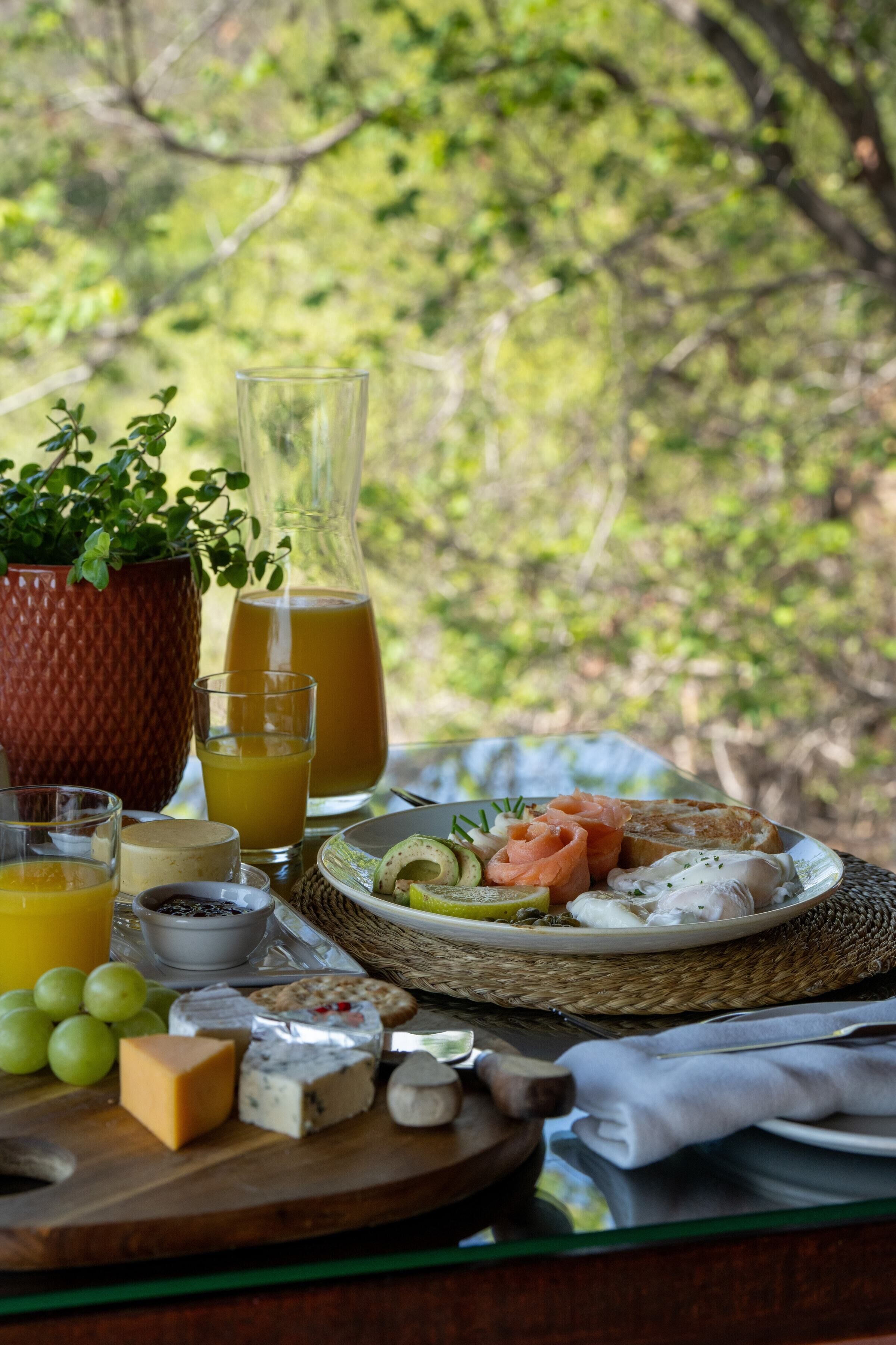 Desayuno con varios quesos, frutas y bebidas sobre una mesa con vista a la vegetación.