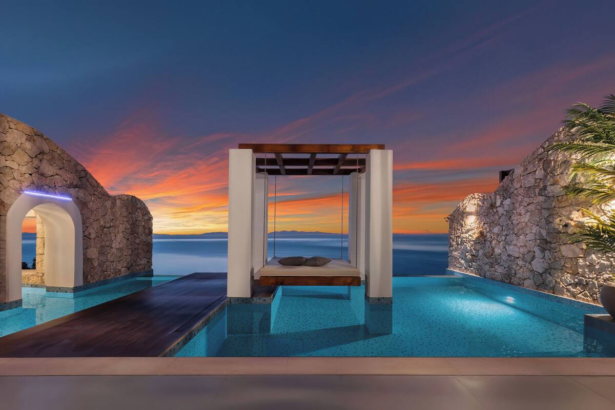 Un espace piscine extérieur paisible, avec un fauteuil suspendu, des murs en pierre et une vue imprenable sur le coucher de soleil.