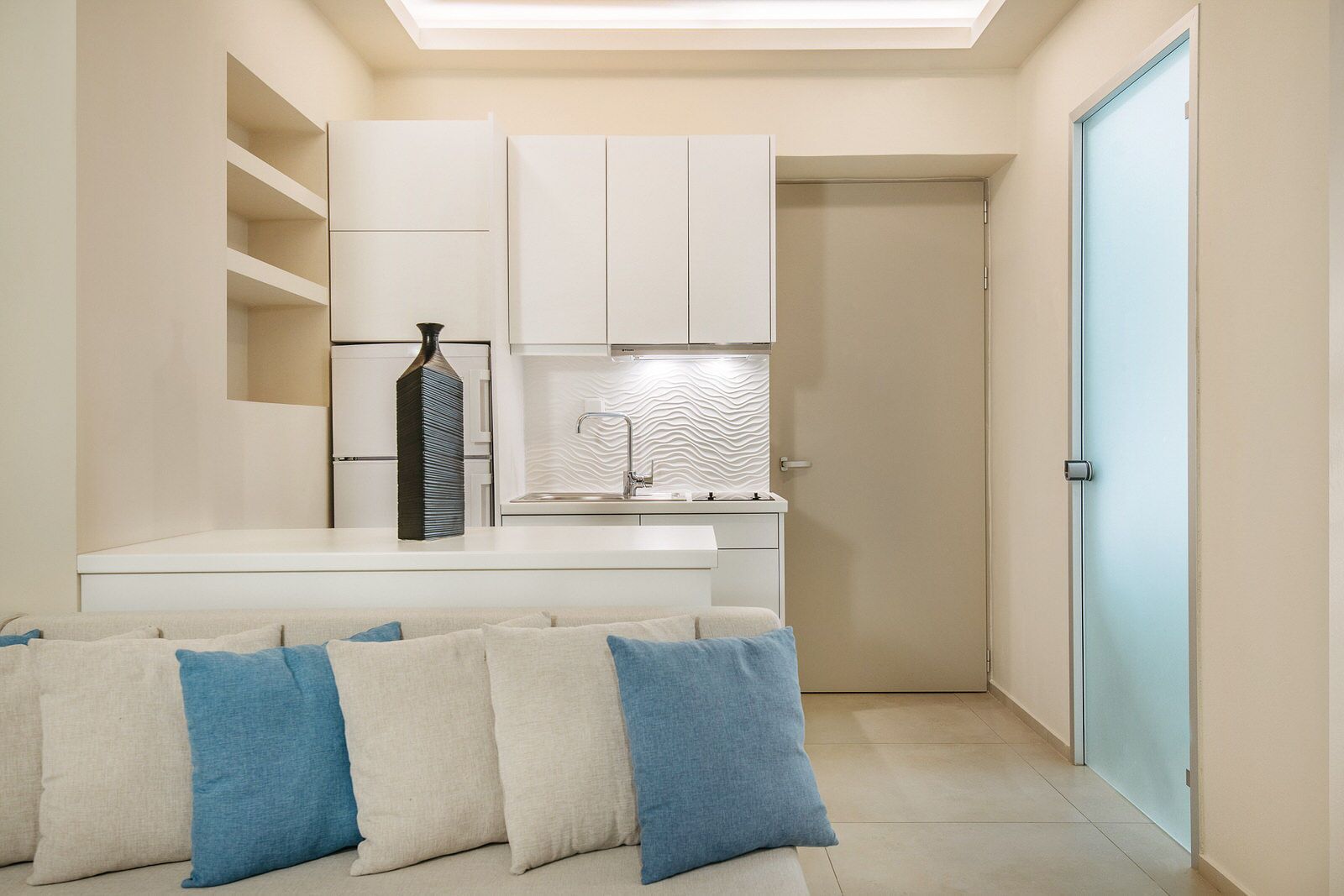 A compact living space with a kitchenette, a sofa with blue cushions, and a door with a frosted glass panel.