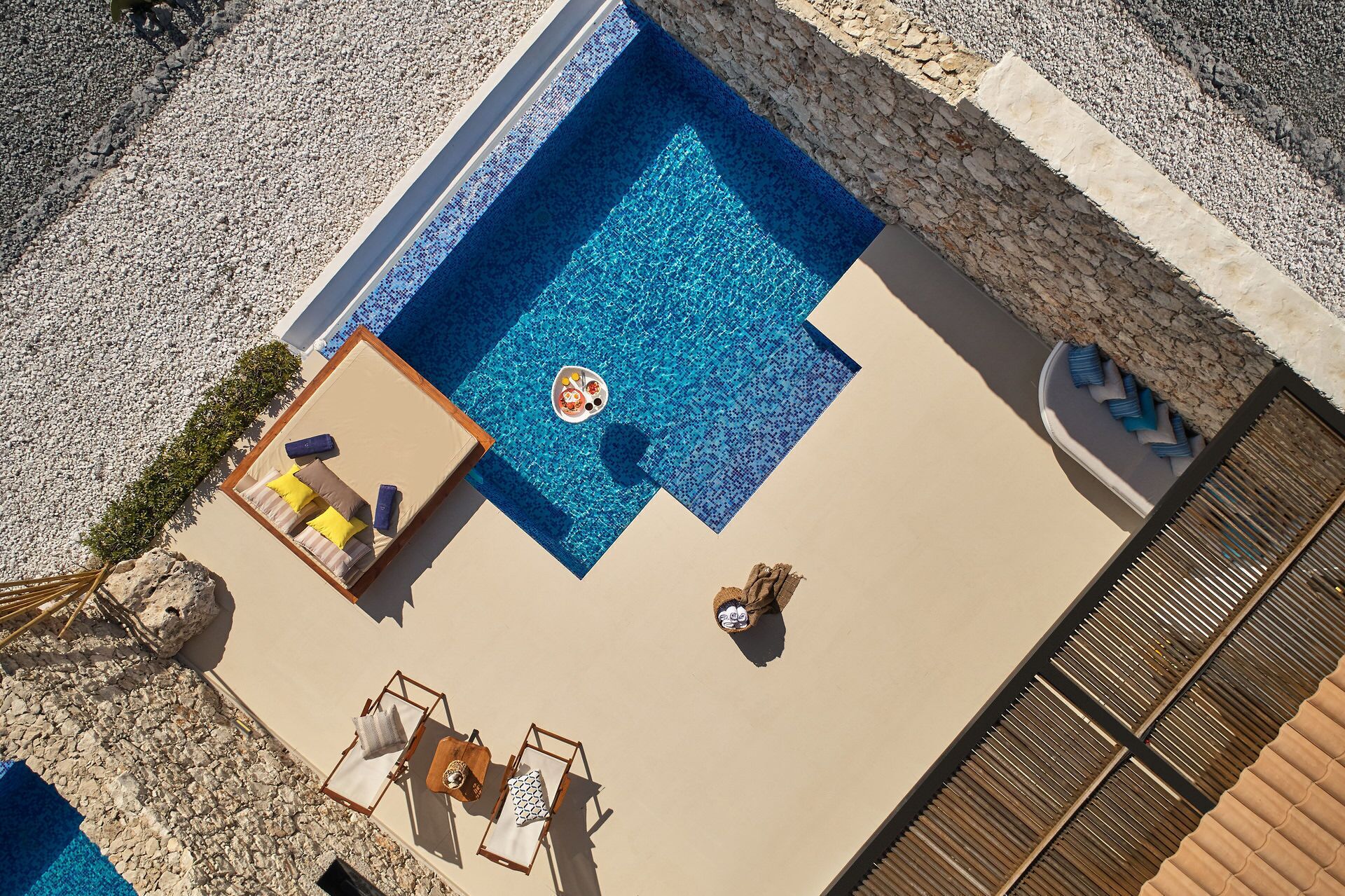 An aerial view of a pool area with a lounge chair and a small table, surrounded by a pebble beach.