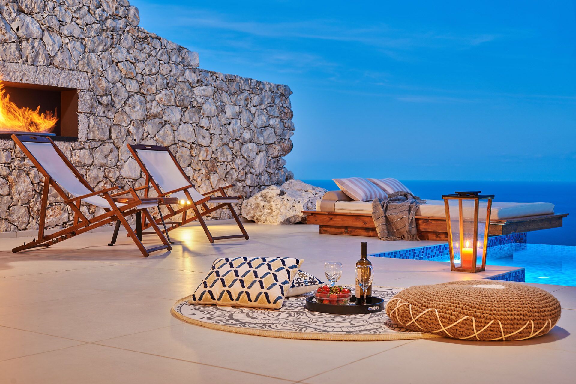 A stone wall, lounge chairs, a sofa, a dining table with food and drink, and a poolside setting with a view of the sea.